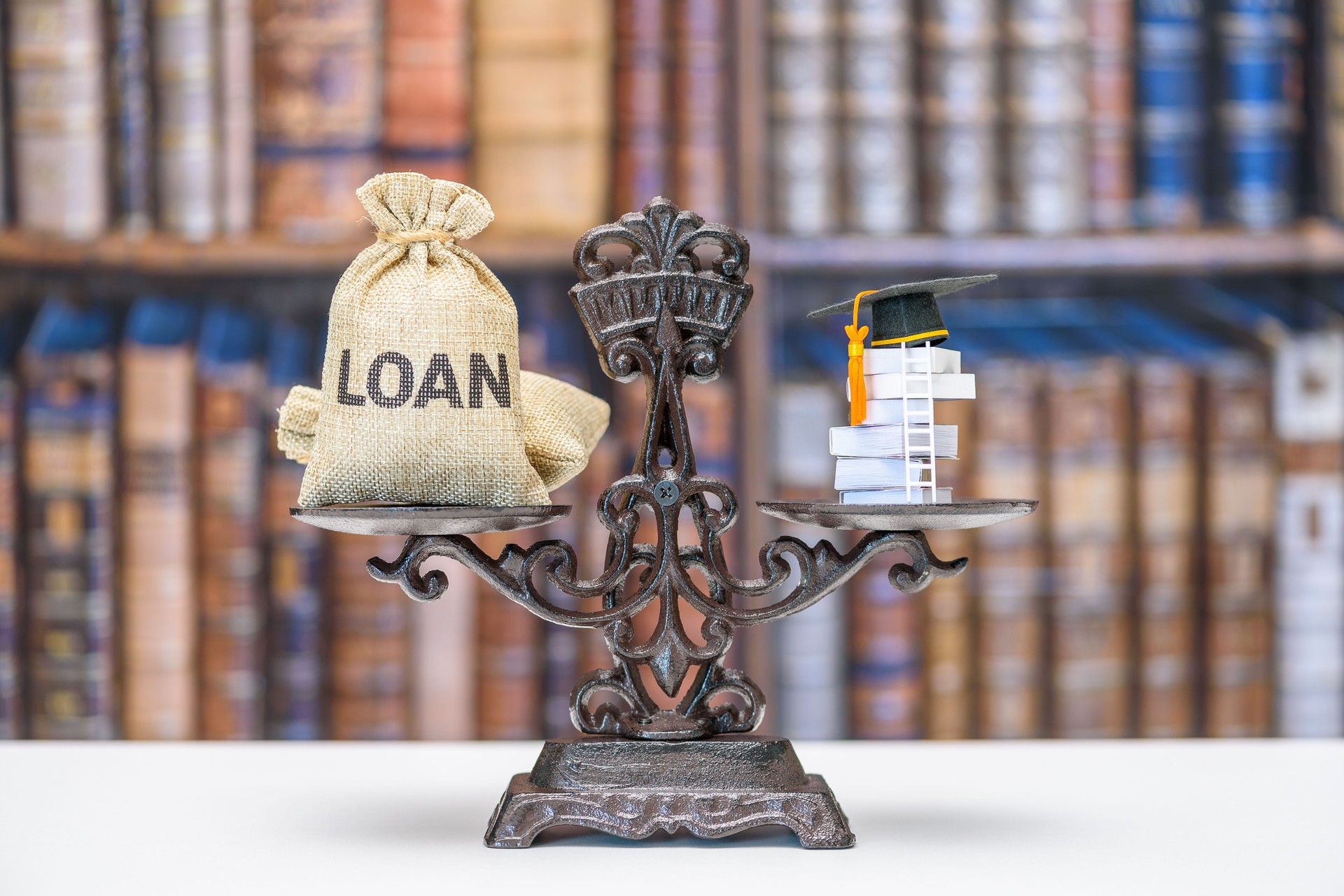 Student loan, education costs, financial burden related to higher education : A balance scale with a sack labeled "LOAN" on one side and a stack of books topped with a graduation cap on the other.
